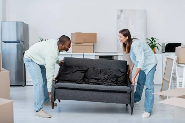 Happy interracial couple carrying sofa in living room — Stock Photo
