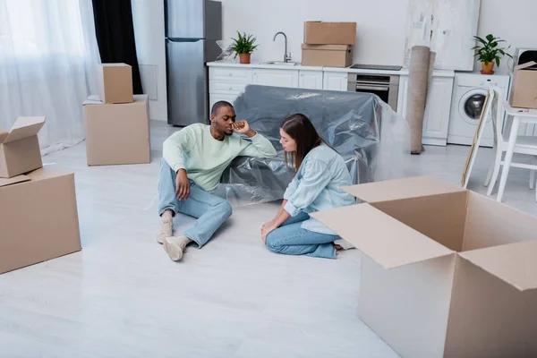 Müdes multiethnisches Paar sitzt auf dem Boden neben Schachteln und Sofa in Plastikfolie — Stockfoto