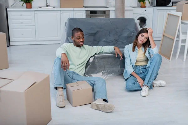 Unzufriedenes multiethnisches Paar sitzt in Plastikfolie neben Kisten und Sofa — Stockfoto