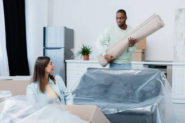 Cheerful woman looking at african american boyfriend holding rolled carpet in new home — Stock Photo