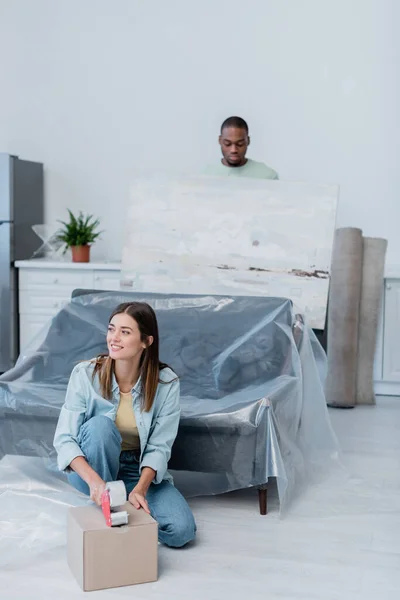Cheerful woman packing box with adjusting tape near african american man holding painting on blurred background — Stock Photo