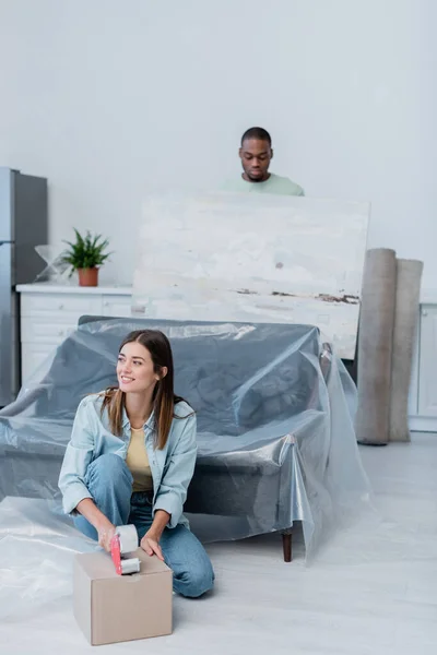 Fröhliche Frau packt Schachtel mit Klebeband in der Nähe afrikanisch-amerikanischer Mann hält Gemälde auf verschwommenem Hintergrund — Stockfoto