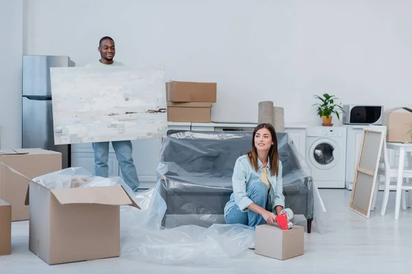 Счастливая молодая женщина упаковки коробку с регулируемой лентой рядом с африканским американским мужчиной проведение живописи в новом доме — стоковое фото