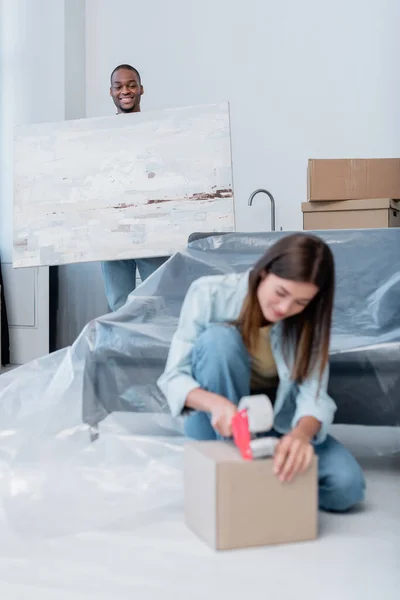 Happy african american man holding painting near blurred young woman packing box with adjusting tape — Stock Photo