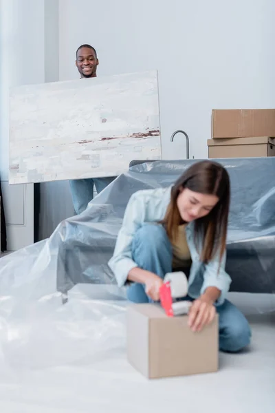 Glücklich afrikanisch-amerikanischer Mann hält Gemälde in der Nähe verschwommene junge Frau Verpackungskiste mit Klebeband — Stockfoto