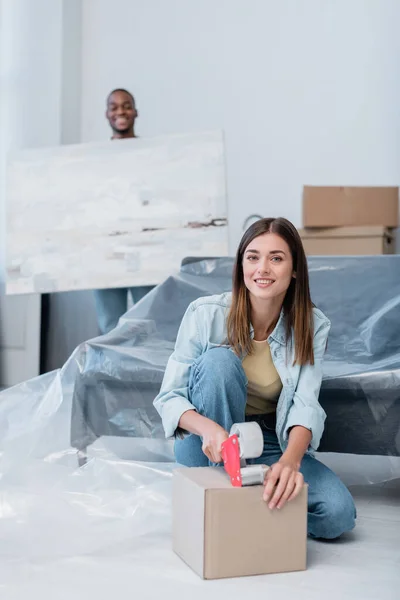 Glückliche junge Frau packt Schachtel mit Klebeband in der Nähe afrikanisch-amerikanischer Mann hält Gemälde auf verschwommenem Hintergrund — Stockfoto
