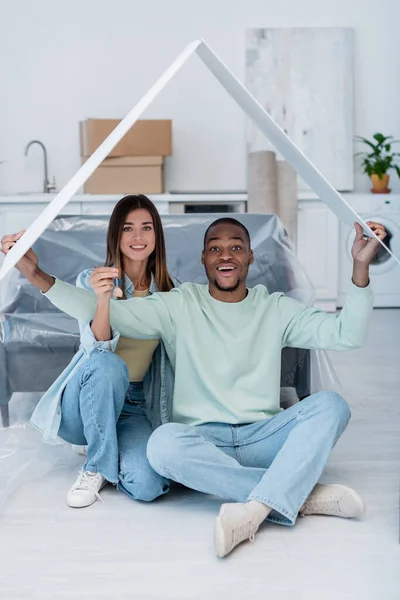 Happy woman holding key while sitting with african american man under paper roof in new apartment — Stock Photo