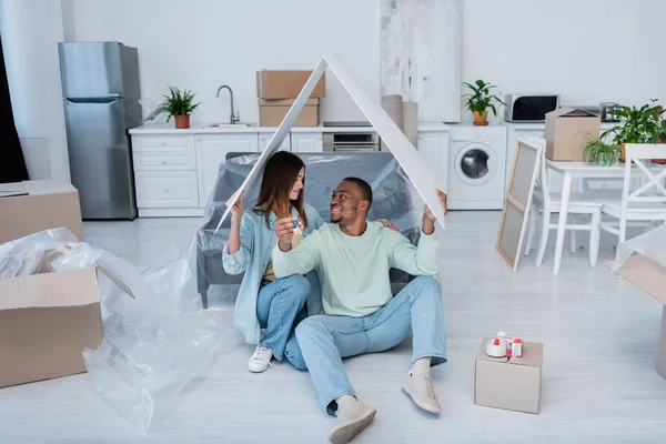 Hombre afroamericano feliz sosteniendo la llave mientras está sentado con su novia bajo el techo de papel en nuevo apartamento - foto de stock