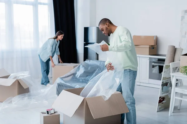 African american man holding plastic wrapper near box and blurred girlfriend during relocation — Stock Photo