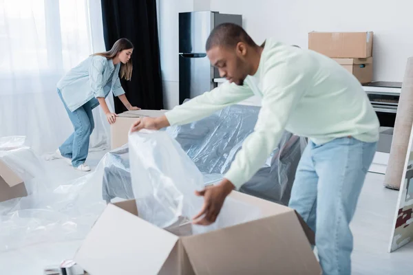 Tätowierte Frau schaut auf Schachtel in der Nähe afrikanisch-amerikanischer Mann mit Plastikverpackung auf verschwommenem Vordergrund — Stockfoto