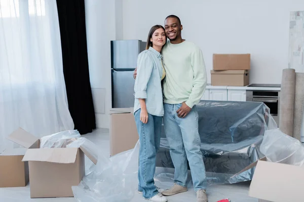 Cheerful interracial couple hugging while standing in new apartment — Stock Photo