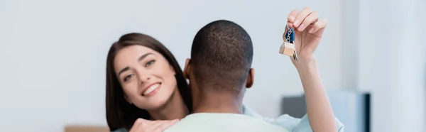 Mujer feliz sosteniendo la llave del nuevo apartamento mientras se abraza con el novio afroamericano, pancarta - foto de stock