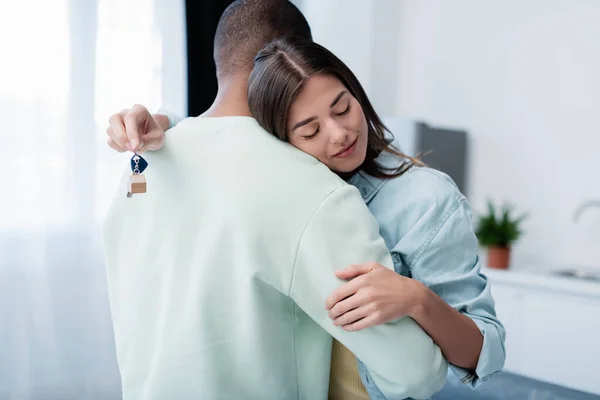 Mujer feliz con los ojos cerrados sosteniendo la llave de nuevo apartamento mientras se abraza con novio afroamericano - foto de stock