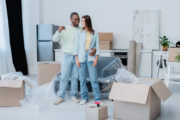 Glücklich afrikanisch-amerikanischer Mann hält Schlüssel, während er fröhliche Freundin in neuer Wohnung umarmt — Stockfoto