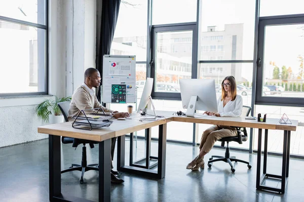 Gente de negocios interracial en gafas mirando monitores de computadora mientras trabaja en la oficina moderna - foto de stock