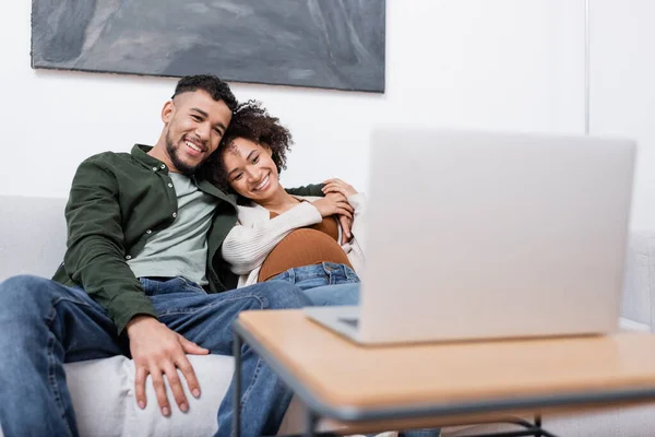 Hombre afroamericano complacido abrazando a la esposa embarazada mientras ve la película en el portátil - foto de stock
