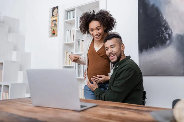 Happy pregnant african american woman pointing at belly near husband during video call on laptop — Stock Photo