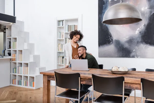 Hombre afroamericano feliz apoyado en el vientre de la esposa embarazada en apartamento moderno - foto de stock