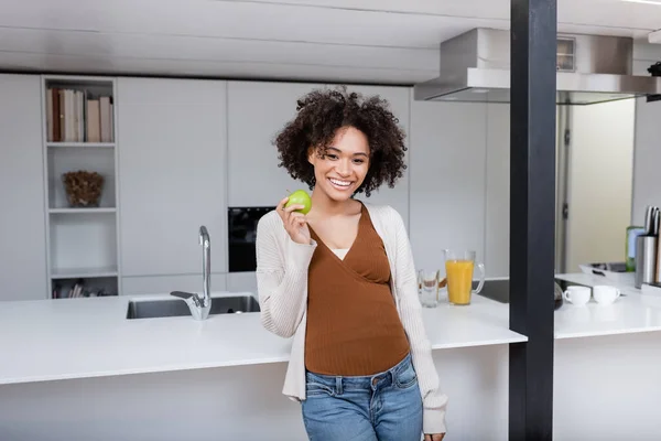 Cheerful pregnant african american woman holding green apple in kitchen — Stock Photo