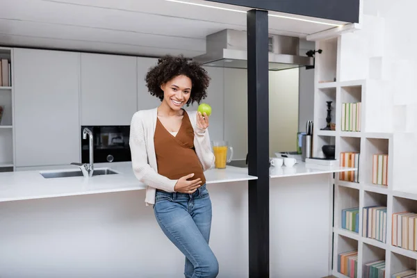 Happy pregnant african american woman holding green apple in kitchen — Stock Photo