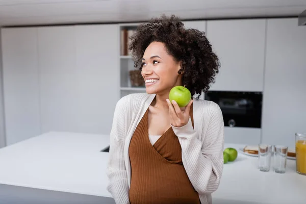 Pleased pregnant african american woman holding green apple in kitchen — Stock Photo