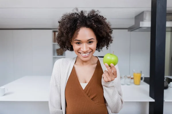 Позитивная и беременная африканская американка держит зеленое яблоко на кухне — стоковое фото