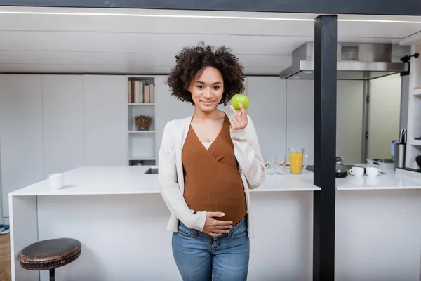 Sorridente donna africana americana incinta che tiene la mela in cucina — Foto stock