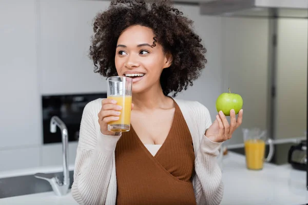 Mulher americana africana grávida alegre segurando maçã verde e vidro de suco de laranja na cozinha — Fotografia de Stock