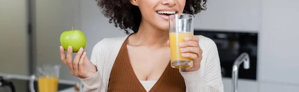 Vista recortada de la alegre mujer afroamericana sosteniendo manzana verde y vaso de jugo de naranja en la cocina, pancarta - foto de stock