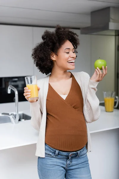 Улыбающаяся беременная американка с зеленым яблоком и стаканом апельсинового сока на кухне — стоковое фото