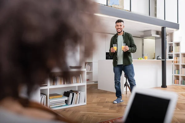 Cheerful african american man holding glasses with fresh orange juice while walking near blurred girlfriend — Stock Photo