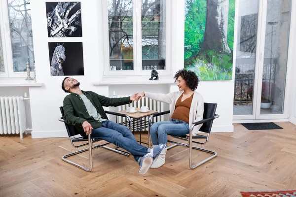 Cheerful african american man laughing while holding hands with happy pregnant wife — Stock Photo