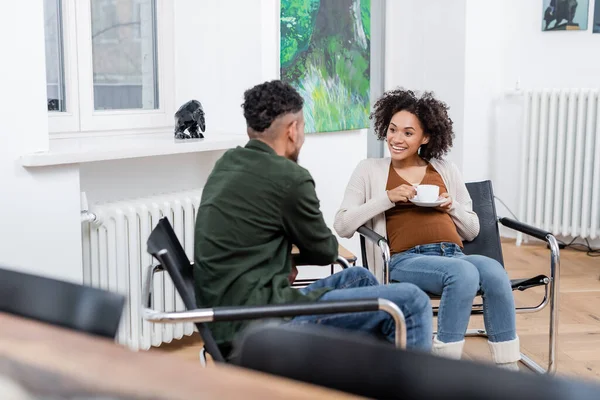 Cheerful african american pregnant woman holding cup and looking at husband at home — Stock Photo