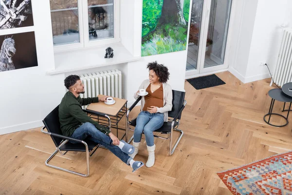 Alto ángulo vista de alegre africana americana embarazada mujer sosteniendo taza y mirando sonriente marido en casa - foto de stock