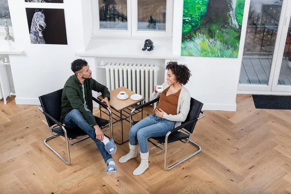 Vista ad alto angolo di allegra donna africana americana incinta in possesso di tazza e guardando il marito riccio a casa — Foto stock