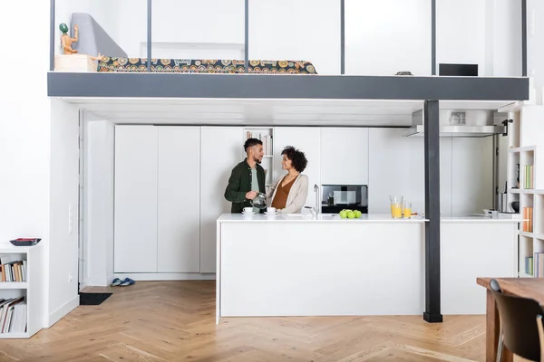 Homem americano africano feliz olhando para a esposa grávida enquanto segurando panela de café na cozinha — Fotografia de Stock