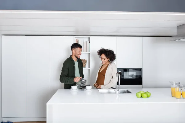 Homme afro-américain joyeux regardant femme enceinte tout en tenant cafetière dans la cuisine — Photo de stock