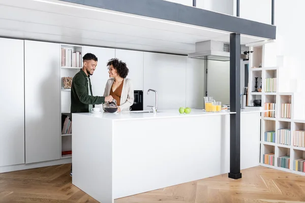 Souriant homme afro-américain regardant femme enceinte tout en tenant cafetière dans la cuisine — Photo de stock