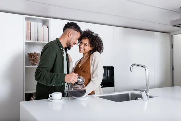 Feliz afro-americano homem derramando café perto alegre grávida esposa — Fotografia de Stock