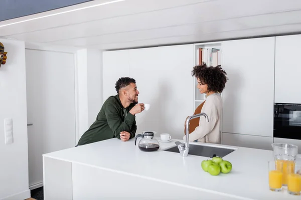 Feliz africano americano hombre mirando embarazada esposa mientras celebración café olla en cocina - foto de stock