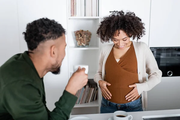 Homme afro-américain flou regardant femme enceinte tout en tenant tasse avec café dans la cuisine — Photo de stock