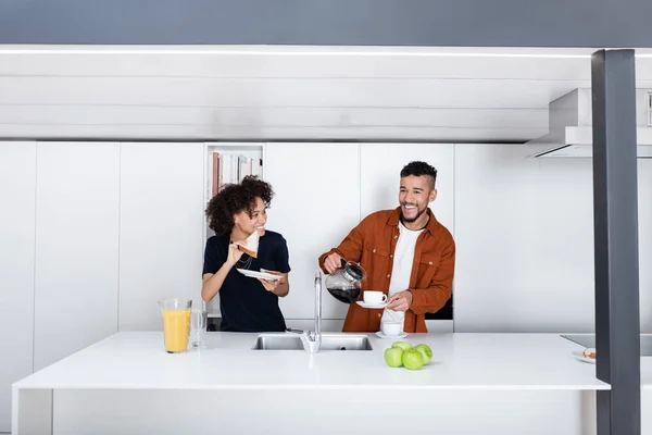 Cheerful african american woman holding sandwich near boyfriend pouring coffee in cup — Stock Photo