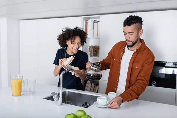 Happy african american woman holding sandwich near boyfriend with coffee pot looking at cups — Stock Photo