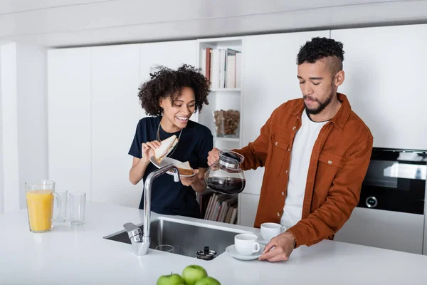 Glücklich afrikanisch-amerikanische Frau hält Sandwich in der Nähe Freund mit Kaffeekanne Blick auf Tassen — Stockfoto