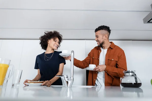 Glücklicher afrikanisch-amerikanischer Mann hält Tassen Kaffee neben Frau in Küche — Stockfoto