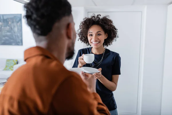 Fröhliche afrikanisch-amerikanische Frau hält eine Tasse Kaffee in der Hand und schaut ihren verschwommenen Freund an — Stockfoto