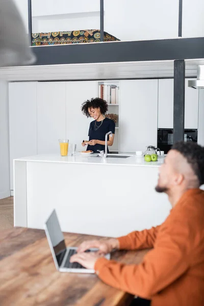 Femme afro-américaine gaie faisant sandwich près de petit ami flou travaillant à la maison — Photo de stock