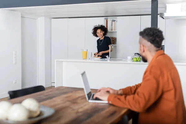 Glücklich afrikanisch-amerikanische Frau macht Sandwich in der Nähe verschwommenen Freund arbeiten aus der Ferne auf Laptop — Stockfoto