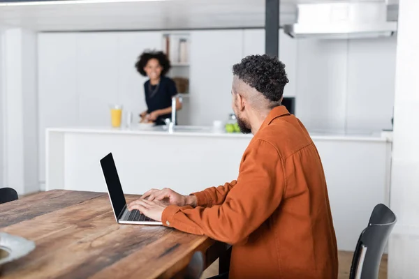 African american man using laptop while working from home and looking at blurred girlfriend in kitchen — Stock Photo