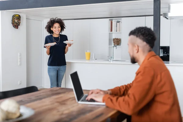 Felice donna afro-americana in possesso di piatti con panini vicino fidanzato offuscata utilizzando il computer portatile mentre si lavora da casa — Foto stock